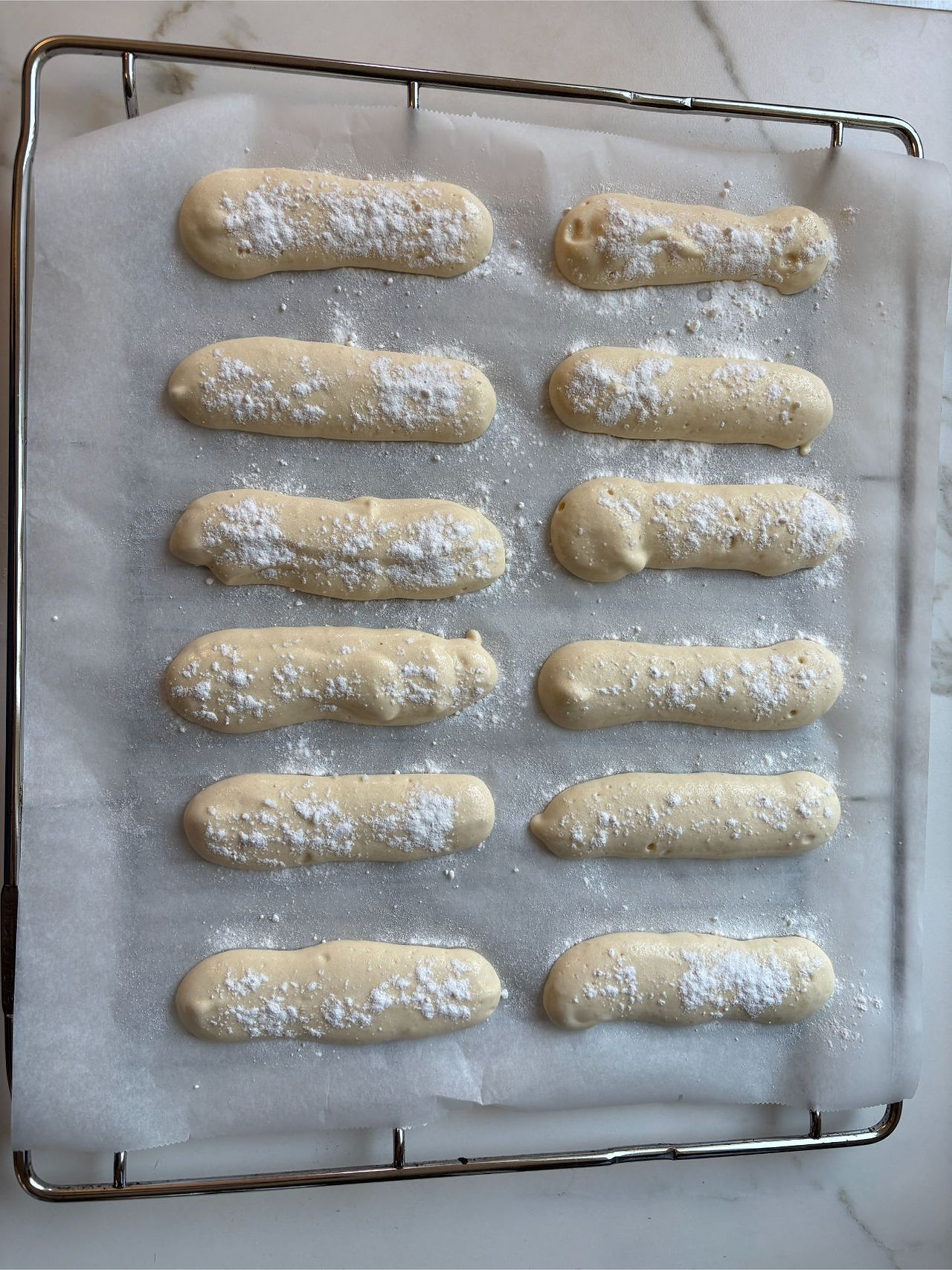 Savoiardi batter being piped into long, narrow fingers on a lined baking tray