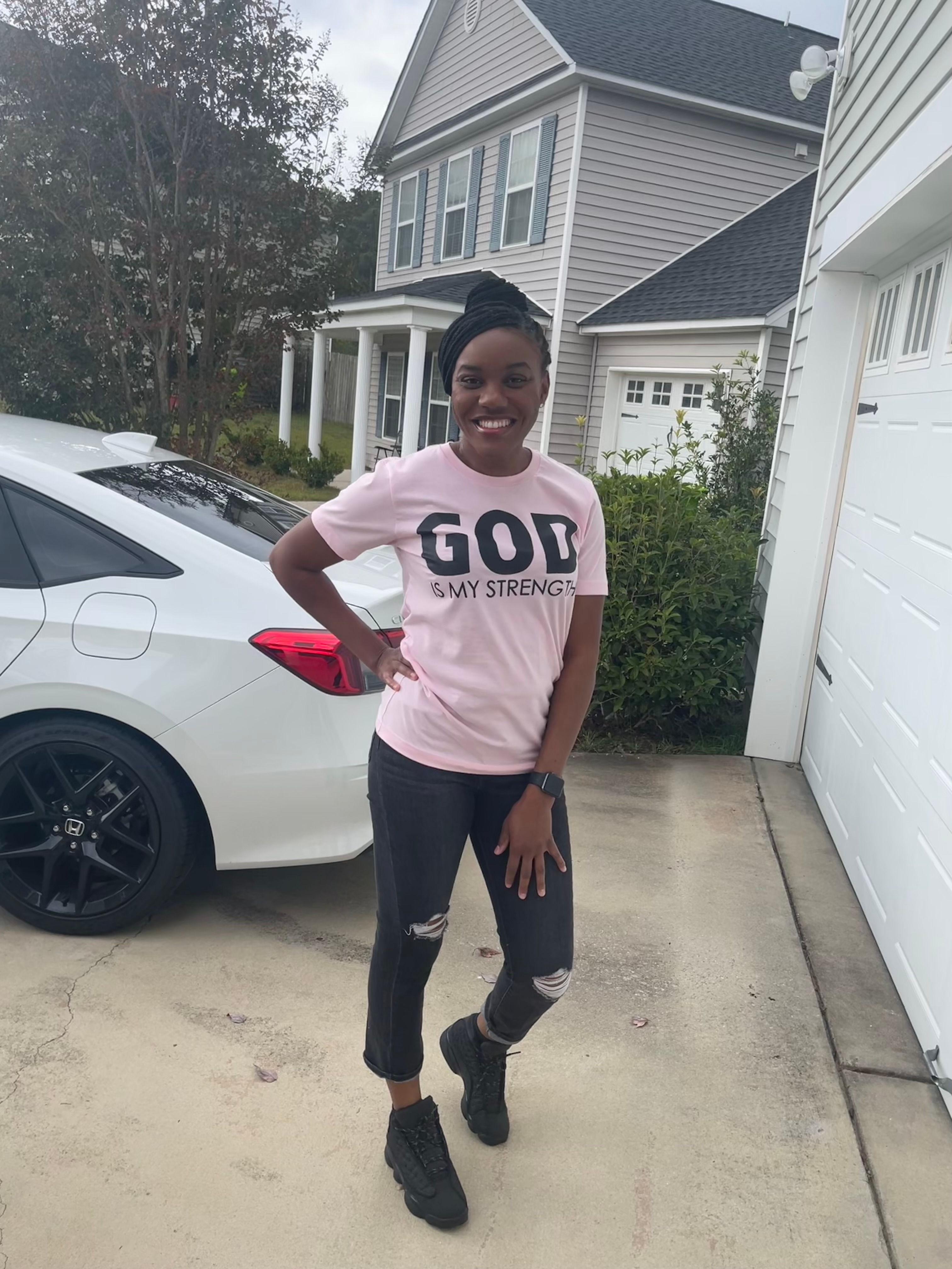 black woman standing in front of white car with pink short sleeve t-shirt with black words God is my strength