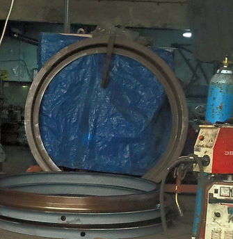 Industrial workshop setting featuring large circular steel components (such as concrete pipe or manhole rings/mould parts), a blue tarp background, and welding or manufacturing equipment on the right.