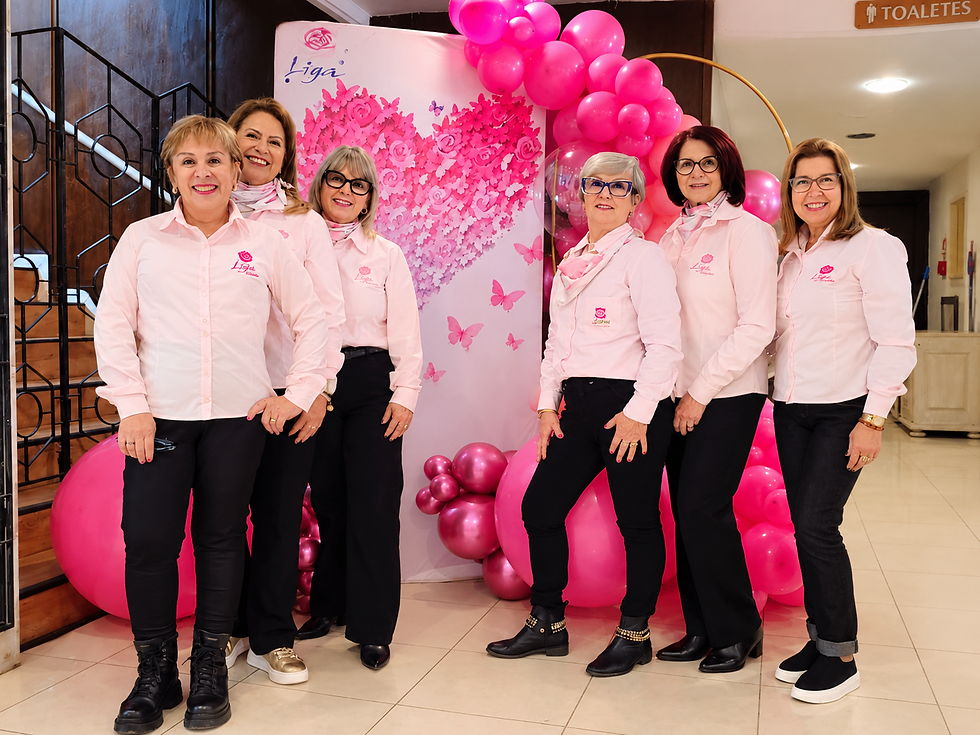 Seis mulheres sorridentes de camisa rosa posam diante de painel com coração e balões rosa; ao fundo,
