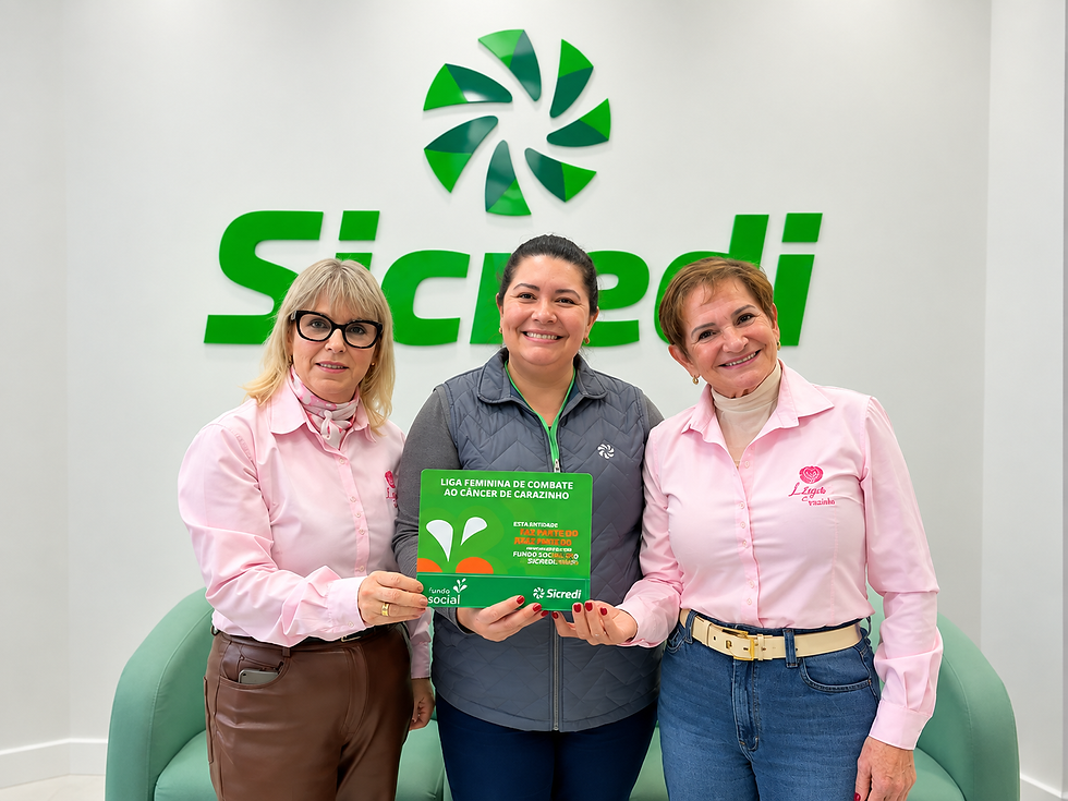 Três mulheres sorrindo em escritório da Sicredi, uma segurando folheto verde da Liga Feminina de Combate ao Câncer.