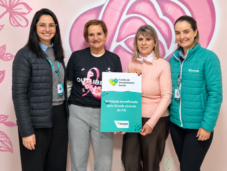 Quatro mulheres sorrindo diante de mural rosa; uma segura placa do Sicoob/FIS com Entidade beneficiada pelo FIS.