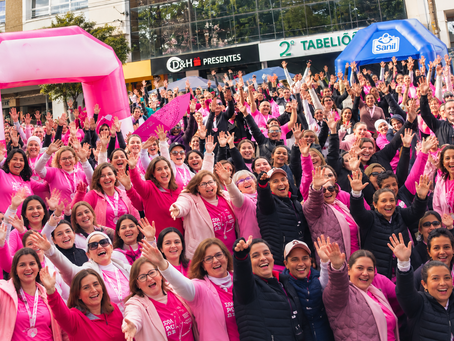 Multidão de mulheres de rosa, sorrindo e erguendo as mãos em evento ao ar livre; faixas D&H Presentes, 2º Tabelião e Sanil.