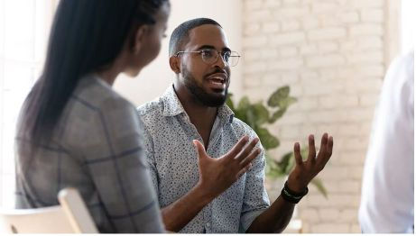 young dark-skinned bearded man wearing glasses uses his hands to speak to a young woman seated beside him