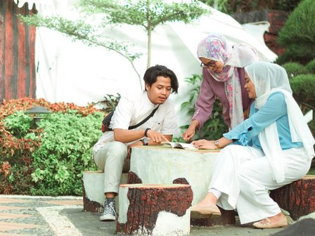 group of 2 muslim women wearing head scarves and a young man sit at a low table in a garden discussing something in a booklet