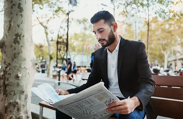 young bearded man in suit sits on park bench next to tree reading newspaper