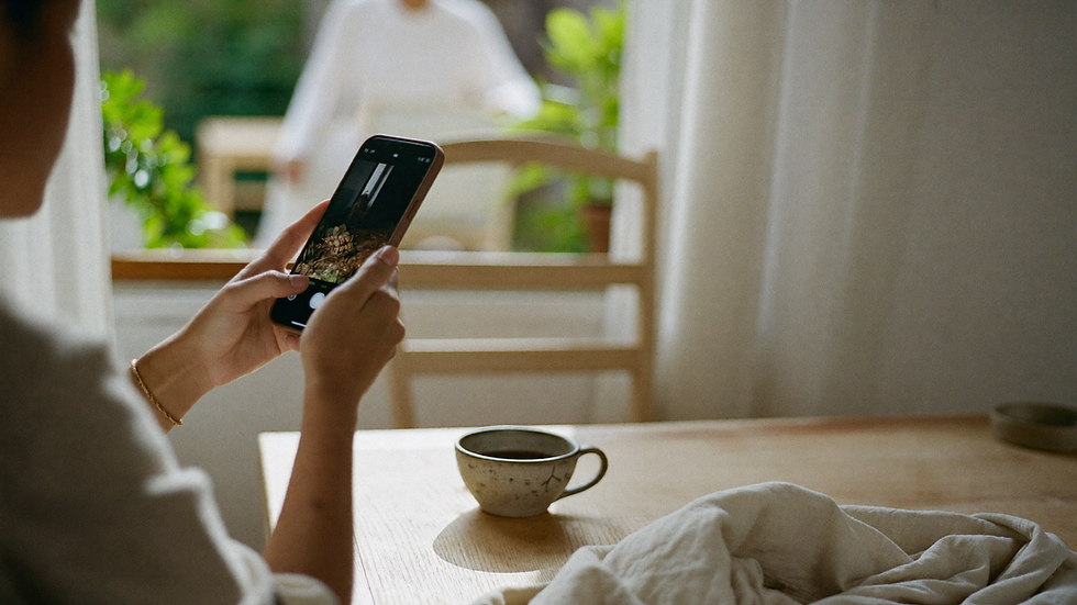 Hands holding a phone at a light wooden table by the window, a ceramic cup of tea and a linen cloth beside them in soft daylight