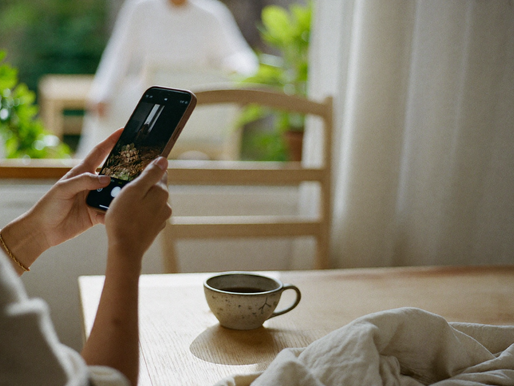Hände halten ein Handy an einem hellen Holztisch am Fenster, daneben eine Keramiktasse Tee und ein Leinentuch im weichen Tageslicht