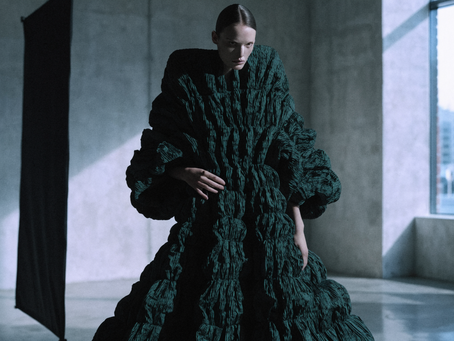 Model in a sculptural dark green gown standing in a concrete studio between two tall lighting flags, lit by cool window light