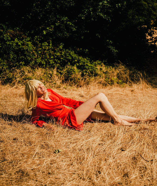 The image captures a confident model showcasing a striking red outfit while sitting on lush grass under bright sunlight.