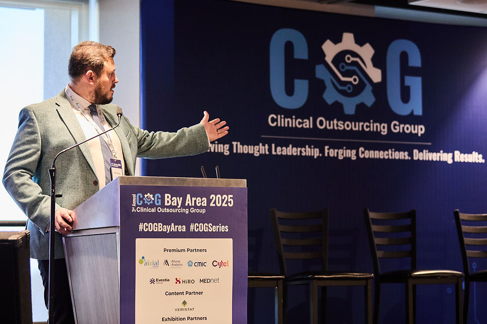 Man in teal blazer speaks at podium, gesturing toward COG Clinical Outsourcing Group backdrop at Bay Area 2025 event.