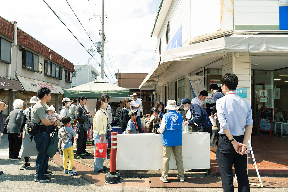青いベストを着たスタッフが屋外イベントでテーブル越しに来場者と交流。周囲には見物する人々が並び、晴れた空が広がる。
