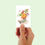 Thumbnail: Hand holding up small card with illustration of a dahlia bouquet against pastel green background.