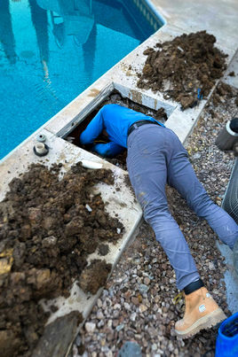 TPB technician leaning their upper body into a hole in the ground next to a pool, likely for a return or skimmer repair.