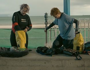 A still from a film - two people are wearing wetsuits and getting ready to go diving.