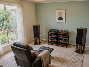 Cozy room with a leather recliner, turntable, and speakers. Soft lighting, green wall, abstract art, window with garden view. Relaxed vibe.