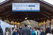 Solstice Lantern Walk banner hanging atop the illuminated Farmers' Market Pavilion entrance, above a crowd entering the market.