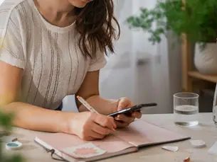A woman is using her phone and planner to create a monthly budget, noting expenses and planning how to manage her finances