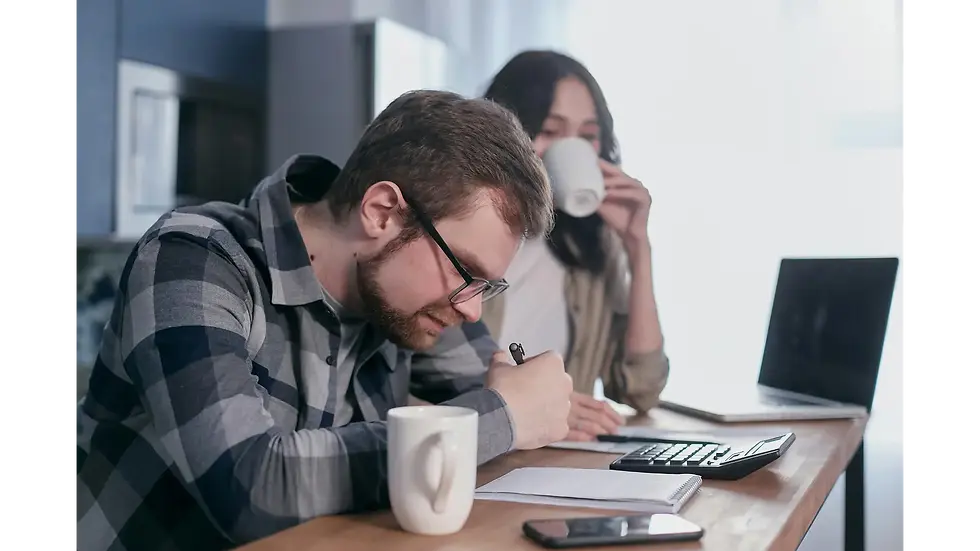 A busy couple budgeting their finances with a calculator, notebook, and laptop at the kitchen table while drinking coffee.