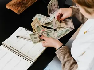 A person counting cash over a desk with a notebook, figuring out how to cut down and budget monthly expenses.
