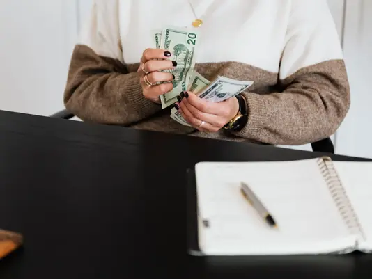 A young woman is counting US dollar bills at a desk with an open notebook, managing her personal budgeting goals.