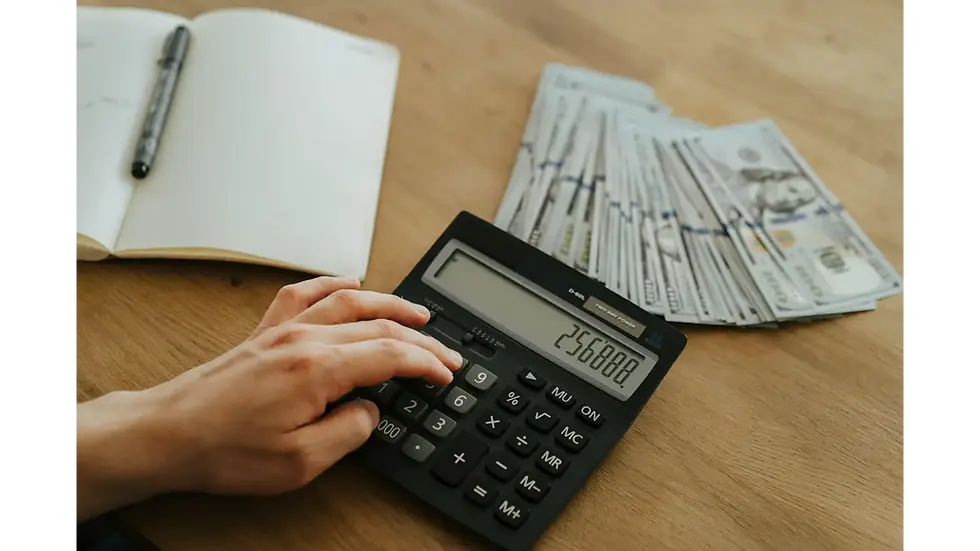 A person using a calculator and cash to manage a paycheck-to-paycheck budget with careful monthly spending plans