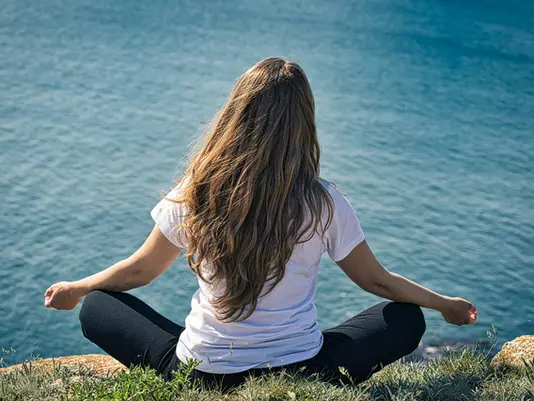 Woman sitting cross-legged in a peaceful meditation pose on a grassy cliff to reduce mind stress.