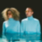 Two people stand in water, wearing matching light blue turtlenecks and sunglasses. Dark background. Calm expression.