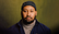 A man with a full beard and a calm, serious expression faces the camera against an olive-green backdrop. He wears a dark beanie, a navy-blue jacket, and a beige turtleneck sweater. A subtle nose ring adds to his distinctive look in this warmly lit portrait.