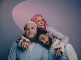 A soft, dreamy portrait of three people posed closely together against a muted purple and blue background with a pink spotlight. A woman with bright pink hair embraces two men—one with a beard and white beanie, wearing a light blue cardigan, and the other with dark hair and a trimmed beard, resting his head gently on the first man's shoulder while wearing a white turtleneck and blue sweater vest. Their pastel-toned clothing and tender expressions create a sense of warmth and connection.
