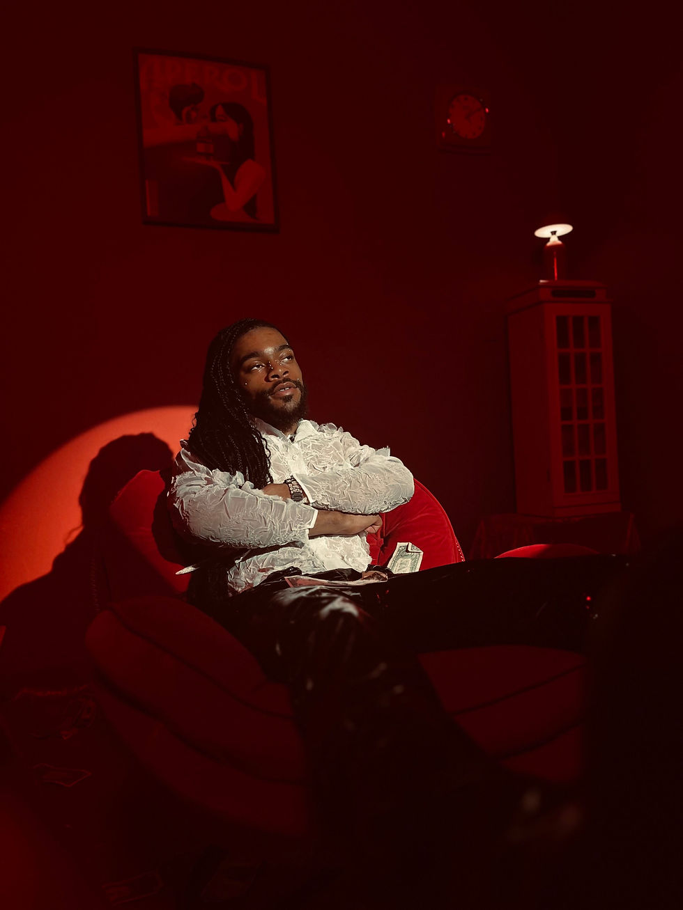 Man in a white shirt sits relaxed in a red-lit room with a lamp and art. Money visible beside him. Mood is calm and contemplative.