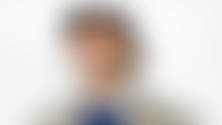 A man with a confident expression looks directly at the camera, wearing bold red eyeshadow that extends dramatically around his eyes. He has short dreadlocks, a mustache, and a visible neck tattoo. Dressed in a textured herringbone coat over a blue sweater and white shirt, he also wears a black baseball cap and a button that says "SEX" in pink letters. The background is a plain white backdrop, emphasizing his striking style and attitude.