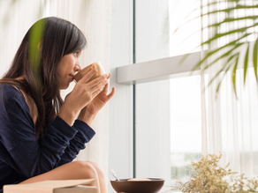 Woman in a blue sweater sipping from a cup by a window. Soft light filters through curtains. Plants add a serene touch to the scene.