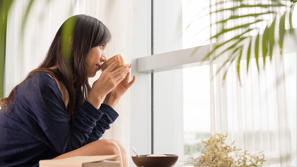 Woman in a blue sweater sipping from a cup by a window. Soft light filters through curtains. Plants add a serene touch to the scene.