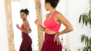 Smiling woman in pink sportswear looks in a mirror while dancing or exercising in a bright room with wood beams and plants