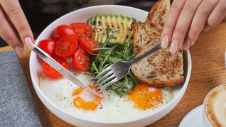Hands with manicured nails hold utensils over a plate of eggs, tomatoes, avocado, arugula, and bread. A cappuccino sits nearby on a wooden table.