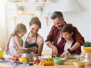 Family cooking in a bright kitchen. A girl reads a cookbook; parents and son prepare vegetables. Smiling, cozy atmosphere.