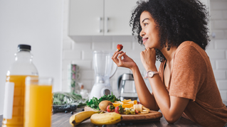Woman in a kitchen smiles while holding a strawberry. Fresh fruits and juice on the table. Bright, cozy atmosphere.
