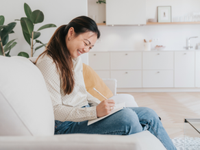 Smiling woman writing in a notebook on a couch in a bright living room with plants, white cabinets, and a glass table with a mug.