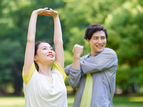 Two people stretching in a park, smiling. One raises arms overhead, the other stretches arm across chest. Lush green trees in background.