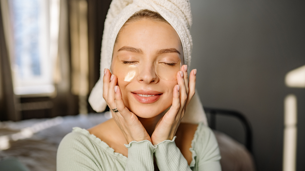 Woman with a towel wrap, eyes closed, smiling while applying skincare patches. Soft lighting, blurred bedroom background, serene mood.