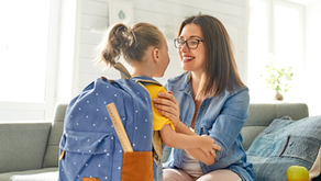 Smiling woman in glasses holds a young girl with a blue backpack on a couch in a bright living room.