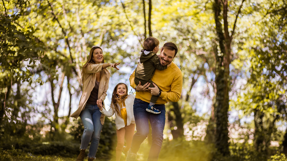 Family joyfully plays in a sunlit forest. A man carries a child; a woman and girl laugh. Greenery surrounds them, creating a cheerful scene.