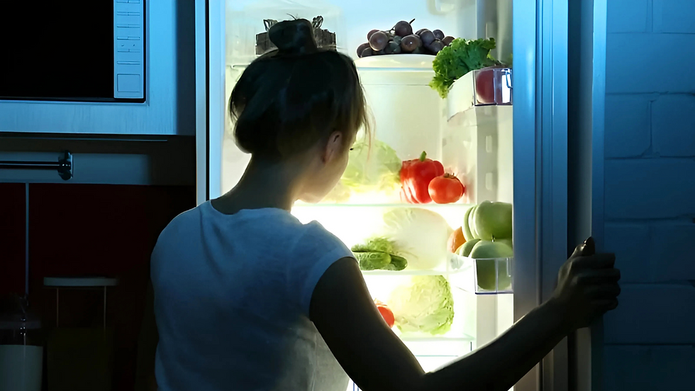 Woman in a white shirt opens a fridge full of fruits and vegetables, casting a warm glow in a dimly lit kitchen.