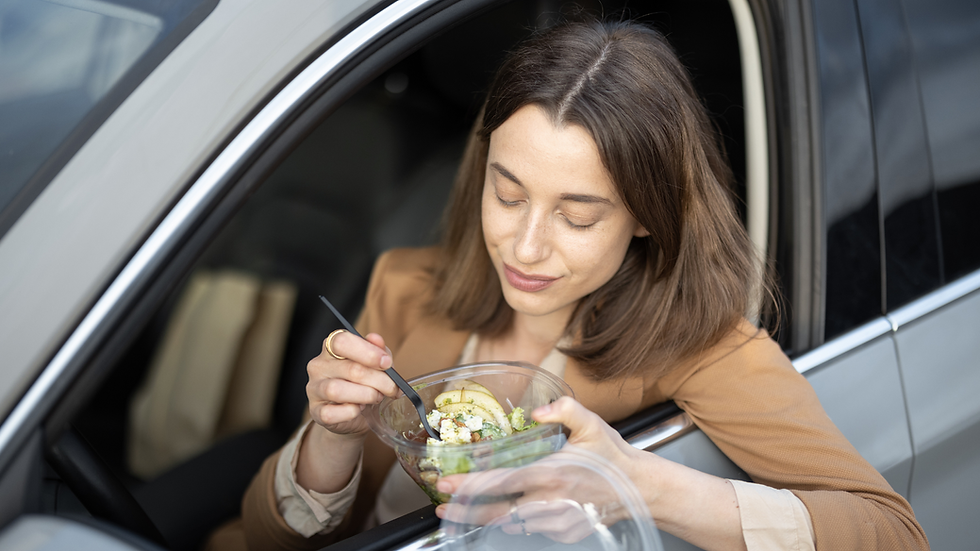 Woman in a car eating a salad with a fork, wearing a brown jacket. The car window is open, and she looks content.