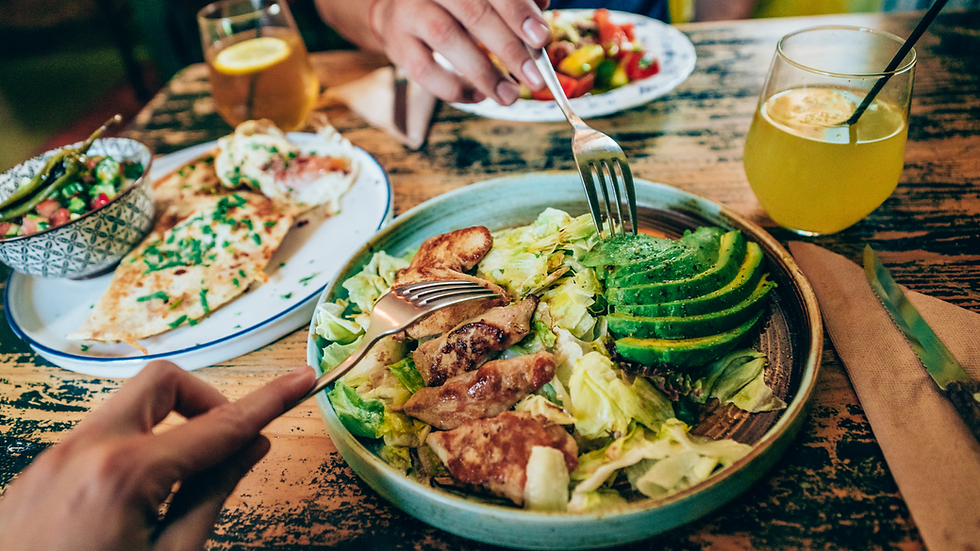 Two hands with forks share a salad with avocado and chicken. Nearby, a quesadilla and a drink with lemon on a rustic wooden table.