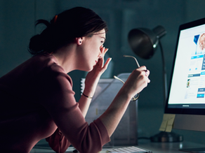 Woman in dim room, rubbing eyes while holding glasses, sits at a desk with a glowing computer screen displaying a webpage. Tired mood.
