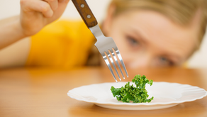 A person in yellow focuses on a fork above a tiny piece of kale on a white plate. The mood is contemplative.