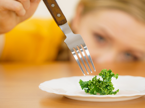 A person in yellow focuses on a fork above a tiny piece of kale on a white plate. The mood is contemplative.