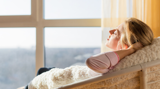 Woman in a cozy white outfit reclines on a dark sofa, reading a book beside a leafy plant in a bright living room.